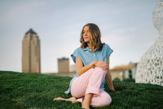 A woman with wavy hair sits barefoot on green grass, wearing a light blue shirt and pink pants, with city buildings and a modern white sculpture visible in the background. by Justin Salem Meyer