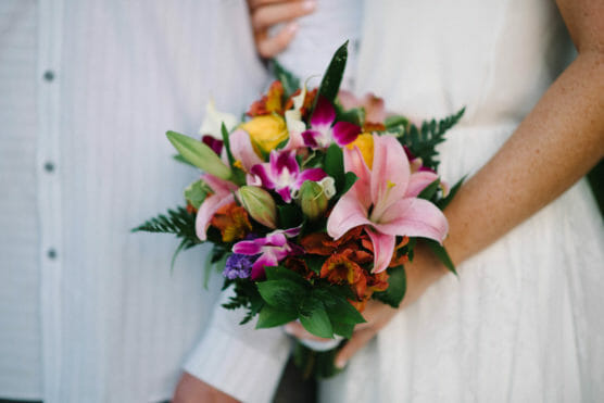 A person in a white dress holds a colorful bouquet of pink lilies, yellow roses, purple orchids, and green ferns, standing beside another person in a white shirt. by Justin Salem Meyer