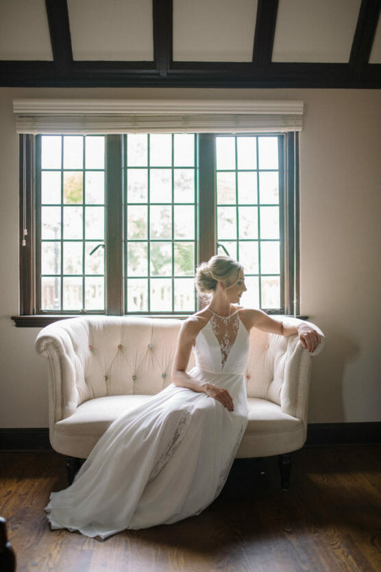 A bride in a white lace gown sits on a cream-colored sofa in front of a large window, looking to the side. Soft natural light fills the elegant room with wooden floors and beamed ceiling. by Justin Salem Meyer