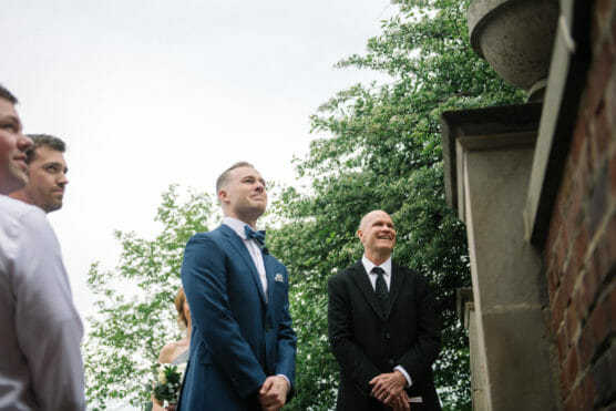 A groom in a blue suit and bow tie stands smiling next to a man in a black suit, outdoors by a brick wall and greenery, with a few other people partially visible. by Justin Salem Meyer