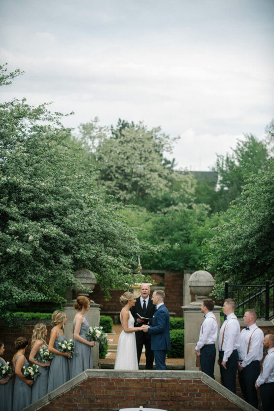 A bride and groom stand facing each other outdoors during their wedding ceremony, surrounded by bridesmaids in gray dresses and groomsmen in white shirts and ties, with lush greenery and trees in the background. by Justin Salem Meyer