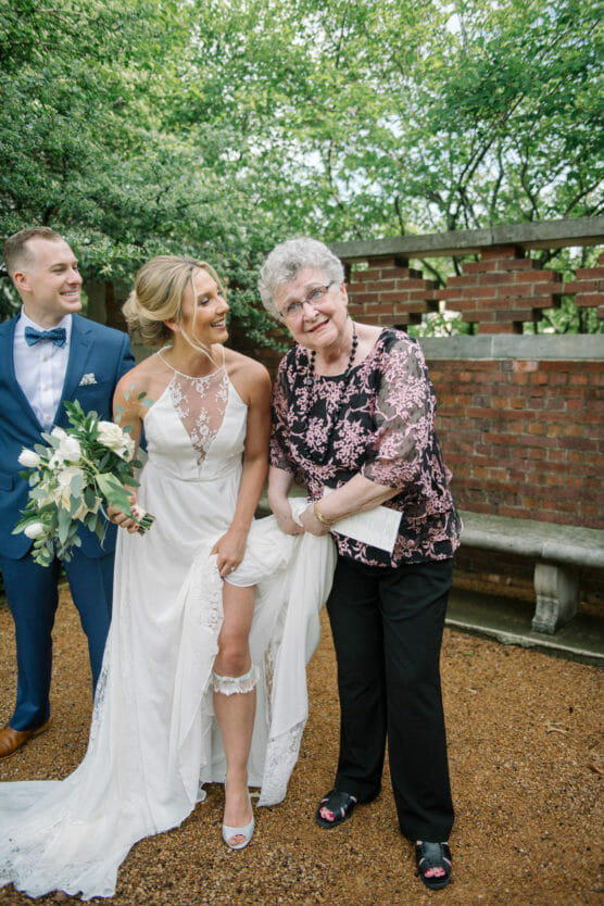 A smiling bride in a white gown playfully lifts her dress beside an older woman in a floral blouse and black pants. A man in a blue suit stands beside them. They are outdoors in a garden with greenery and a brick wall. by Justin Salem Meyer