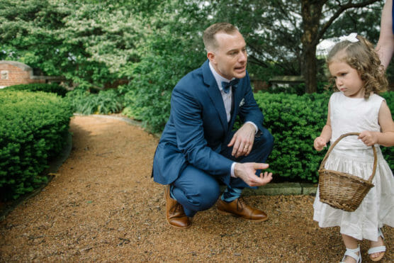 A man in a blue suit crouches on a garden path, talking to a young girl in a white dress holding a wicker basket. The scene is outdoors with green bushes and trees in the background. by Justin Salem Meyer