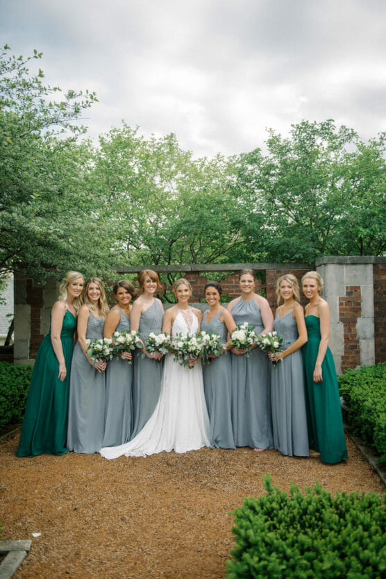 A bride in a white dress stands outdoors with eight bridesmaids in matching gray and green dresses, all holding bouquets, with green trees and a brick wall in the background. by Justin Salem Meyer