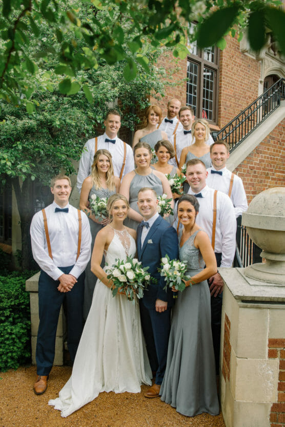 A wedding party poses outdoors by a brick building and stone steps. The bride and groom stand in front, surrounded by bridesmaids in gray dresses and groomsmen in white shirts with suspenders. Everyone is smiling at the camera. by Justin Salem Meyer