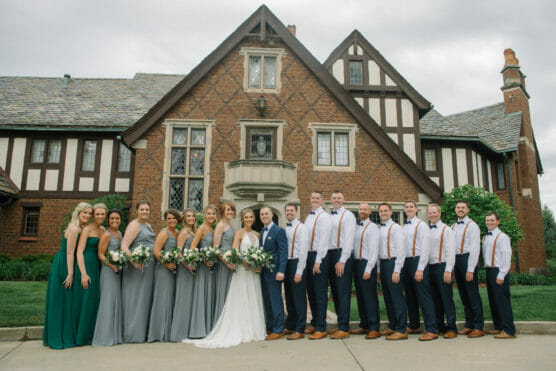 A large wedding party stands in front of a Tudor-style brick house. The bride and groom are in the center, surrounded by bridesmaids in green and gray dresses and groomsmen in white shirts with suspenders and ties. by Justin Salem Meyer