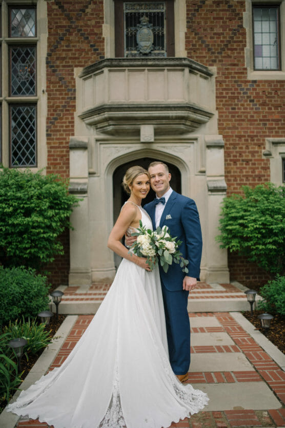 A bride in a white gown and a groom in a navy suit smile as they pose together outside a brick building with greenery and a decorative entrance behind them. by Justin Salem Meyer