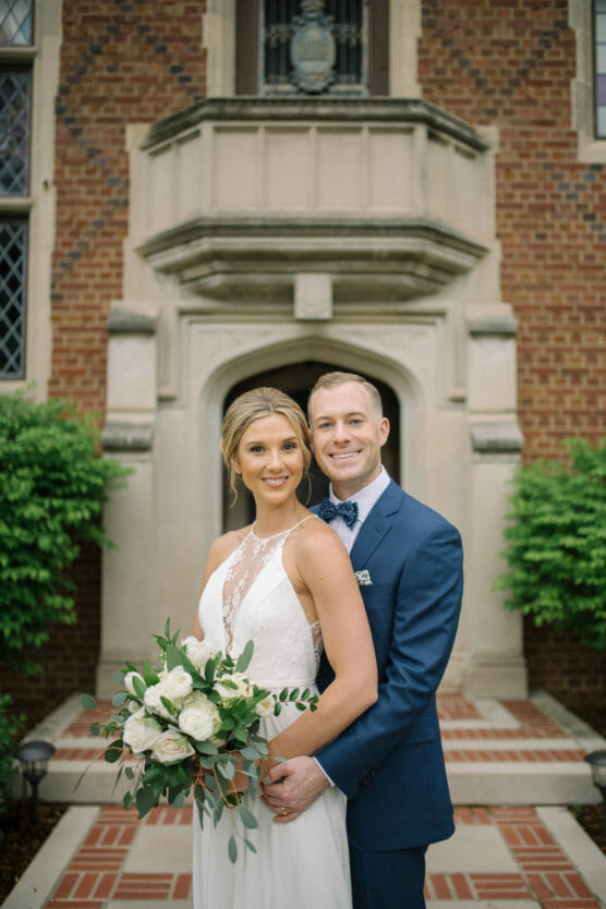 A bride in a white dress holding a bouquet of white flowers stands in front of a groom in a blue suit. They are smiling and posing outside a brick building with greenery and a stone archway. by Justin Salem Meyer