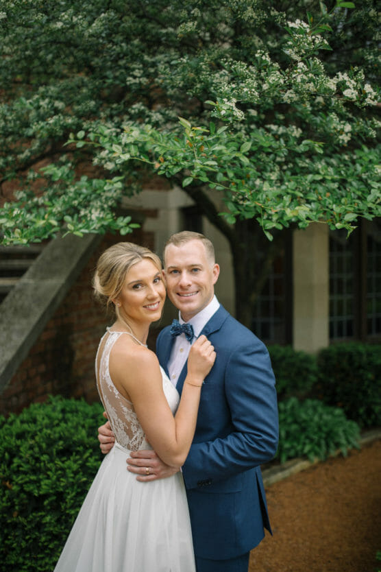 A bride in a white lace dress and a groom in a blue suit embrace and smile at the camera, standing in a garden with lush greenery and a flowering tree in the background. by Justin Salem Meyer
