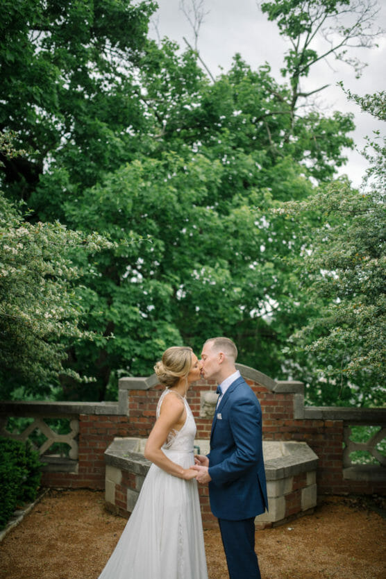 A bride and groom stand outdoors, holding hands and sharing a kiss in front of a stone bench and brick wall, surrounded by lush green trees and foliage. by Justin Salem Meyer