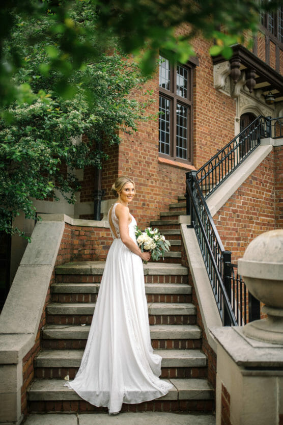 A bride in a white dress stands on outdoor stone steps, smiling over her shoulder and holding a bouquet. Green leaves frame the scene, and a brick building with large windows is in the background. by Justin Salem Meyer