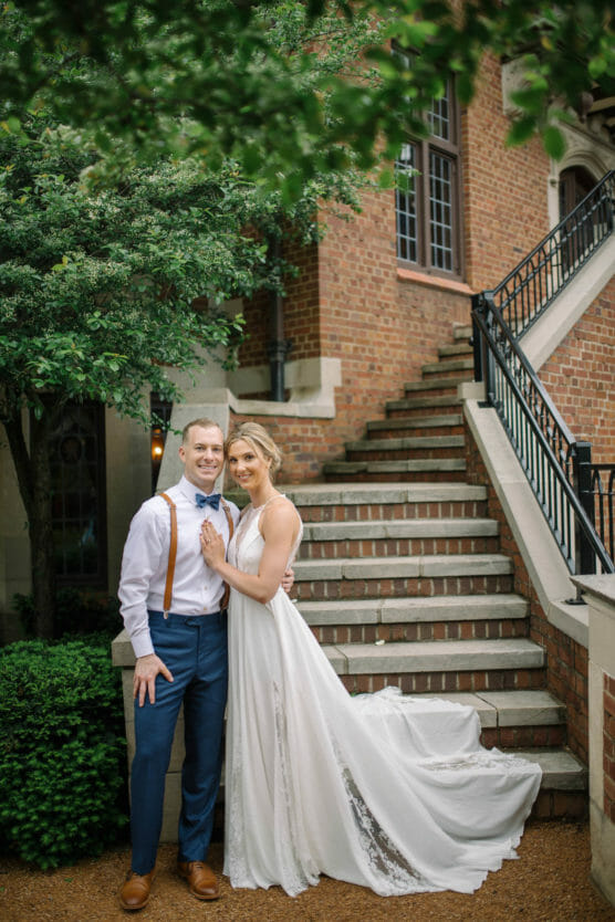A smiling couple poses together outdoors; the woman wears a white wedding dress and the man wears blue trousers, a white shirt, bow tie, and suspenders, standing by a brick staircase and green trees. by Justin Salem Meyer