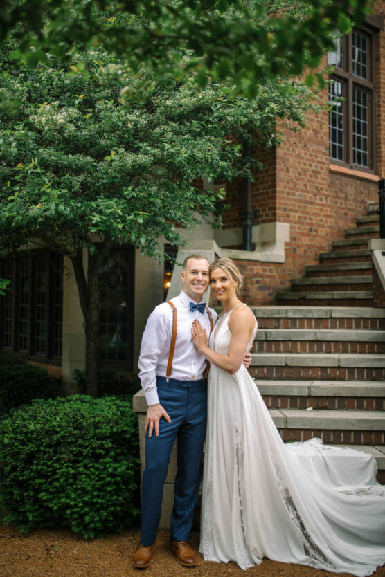 A bride in a flowing white dress and a groom in blue pants, white shirt, and suspenders stand smiling together outside by greenery and brick steps, with trees and a brick building in the background. by Justin Salem Meyer