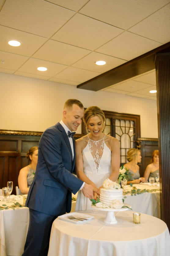 A bride and groom smile as they cut their wedding cake together. The bride wears a white dress with lace details, and the groom is in a blue suit. Bridesmaids are seated at a table in the background. by Justin Salem Meyer