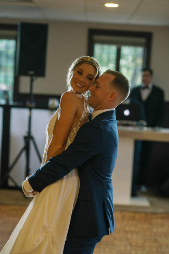 A smiling bride in a white dress and a groom in a blue suit share a joyful dance in a warmly lit room, with a DJ and speakers visible in the background. by Justin Salem Meyer