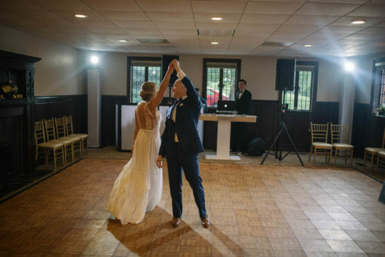 A bride in a white dress and a groom in a navy suit dance together in a mostly empty room, while a DJ stands behind a table in the background. The couple is smiling and holding hands as they twirl. by Justin Salem Meyer