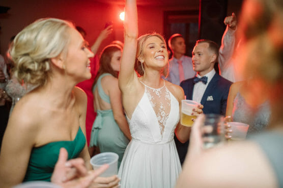 A bride in a white dress smiles joyfully with her arm raised, holding a drink, surrounded by friends dancing and celebrating at a wedding reception. by Justin Salem Meyer