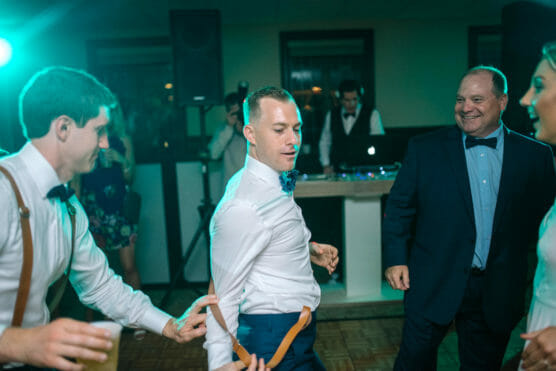 A group of people in formal attire dance energetically at a party, with one man pulling on another mans suspenders. A DJ is seen in the background behind a booth. The atmosphere is joyful and lively. by Justin Salem Meyer