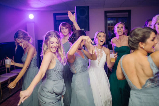 A group of women, including a bride in a white dress, dance and laugh together at a celebration. The atmosphere is lively, with colorful lighting and everyone appearing joyful and energetic. by Justin Salem Meyer