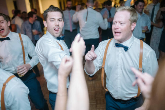 A group of men wearing white shirts, suspenders, and bow ties dance energetically at a party. Two men in the foreground gesture and make playful faces, while others enjoy the lively atmosphere in the background. by Justin Salem Meyer