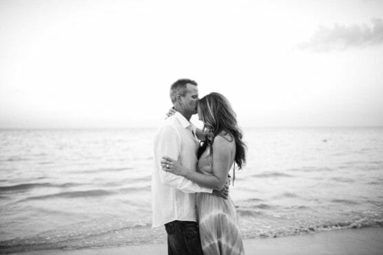 A couple stands close together on a beach, kissing as they embrace. The scene is in black and white, with calm ocean waves and a clear sky in the background. by Justin Salem Meyer