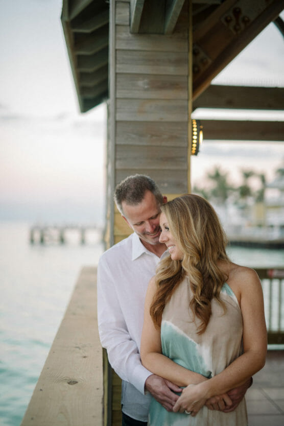 A couple stands on a wooden deck by the water, smiling and embracing. The man stands behind the woman, wrapping his arms around her as she looks ahead. Palm trees and a pier are visible in the soft, scenic background. by Justin Salem Meyer