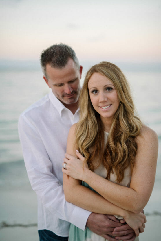 A man and woman stand close together on a beach at sunset. The woman with long blonde hair smiles at the camera while the man stands behind her, holding her gently and looking down. The ocean is calm in the background. by Justin Salem Meyer