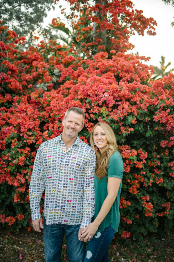 A couple stands holding hands in front of a lush bush covered in bright red-orange flowers. The man wears a patterned button-down shirt and jeans, while the woman wears a green top and ripped jeans. Both are smiling at the camera. by Justin Salem Meyer
