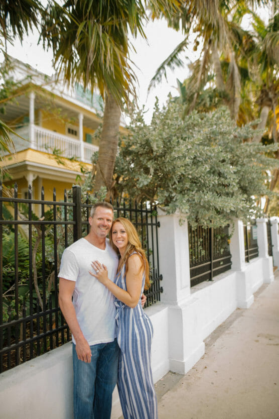 A couple stands smiling together on a sidewalk next to a black iron fence with palm trees and a yellow house in the background. The woman wears a blue striped jumpsuit, and the man wears a white t-shirt and jeans. by Justin Salem Meyer