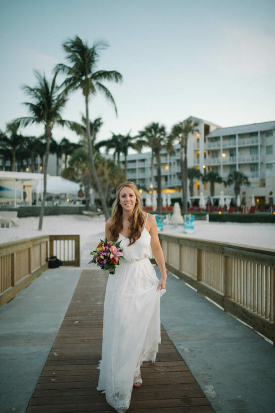A woman in a white dress holds a colorful bouquet while walking on a wooden boardwalk at a beach resort, with palm trees and a hotel in the background. by Justin Salem Meyer