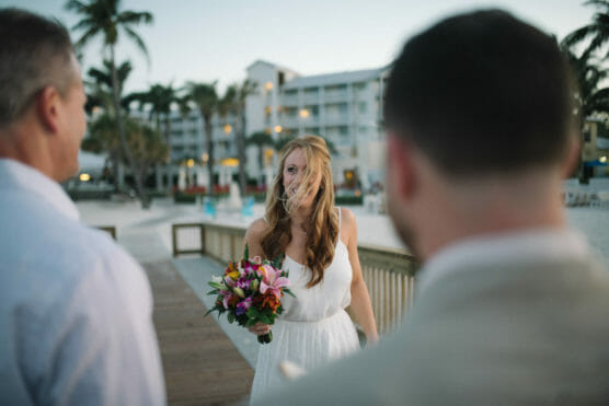 A bride holding a colorful bouquet stands on a boardwalk at a beach resort, her hair windswept across her face. Two people, seen from behind, face her. Palm trees and a multi-story building are in the background. by Justin Salem Meyer
