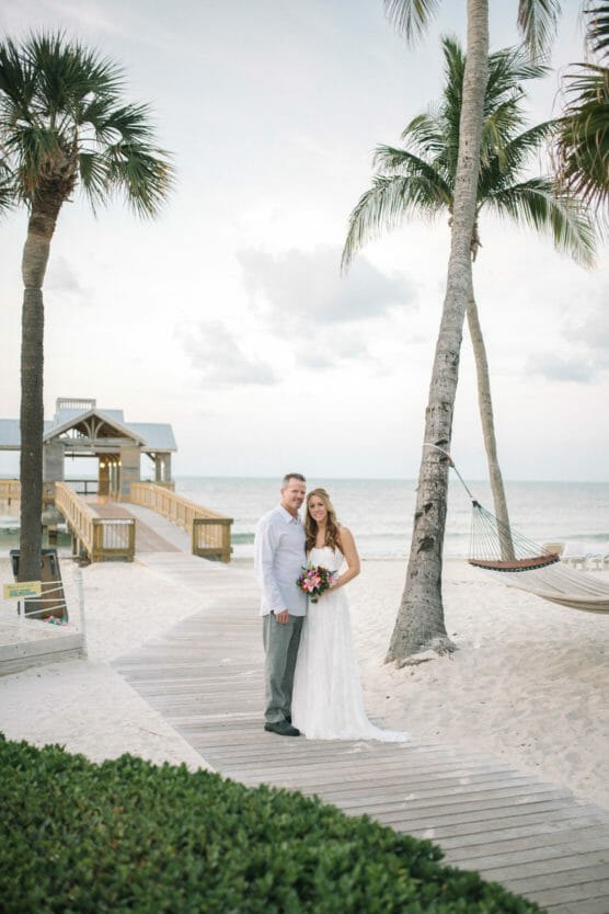 A bride and groom stand together on a wooden pathway at a tropical beach, surrounded by palm trees. The bride holds a bouquet, and a hammock hangs nearby. A small pier extends into the ocean in the background. by Justin Salem Meyer