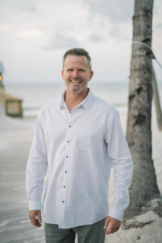 A man with short hair and a trimmed beard, wearing a white button-up shirt, stands and smiles on a beach near a palm tree, with the ocean and boardwalk in the background. by Justin Salem Meyer