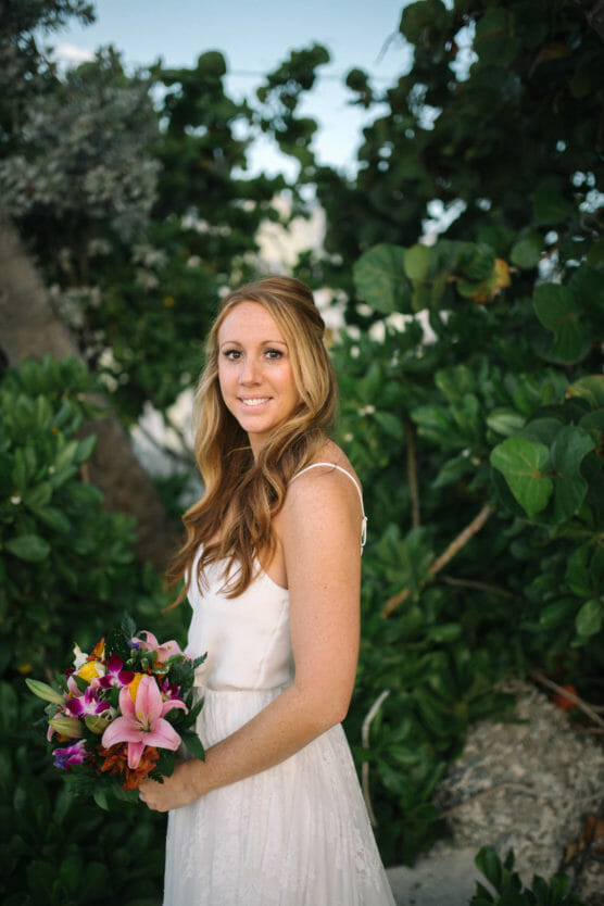 A woman in a white dress stands outdoors holding a colorful bouquet of flowers, surrounded by lush green foliage. She has long, wavy hair and is looking at the camera with a gentle smile. by Justin Salem Meyer
