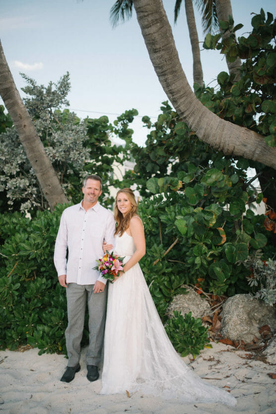 A bride in a white dress holding a bouquet stands next to a man in a white shirt and gray pants on a sandy beach, surrounded by lush green plants and palm trees. by Justin Salem Meyer