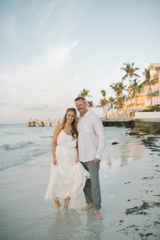 A smiling couple stands barefoot on a sandy beach at sunset, with palm trees, ocean waves, and buildings in the background. The woman wears a white dress and the man wears a light shirt and pants. by Justin Salem Meyer