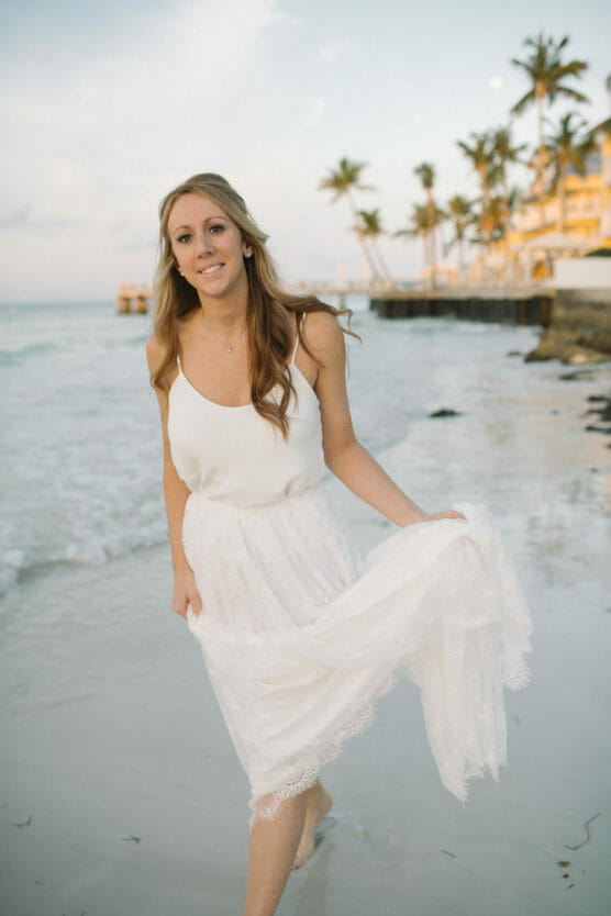 A woman in a white dress walks barefoot along a sandy beach, holding up her skirt slightly. Palm trees and buildings line the shore in the background under a partly cloudy sky. by Justin Salem Meyer