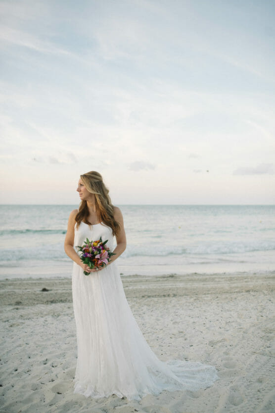 A bride in a flowing white dress holds a bouquet of flowers and stands on a sandy beach, looking to her left, with the ocean and a pastel sky in the background. by Justin Salem Meyer