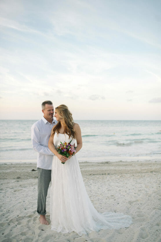 A bride in a white dress holding a bouquet stands on the beach with a groom in a white shirt and gray pants. They smile at each other, with the ocean and a pastel sky in the background. by Justin Salem Meyer