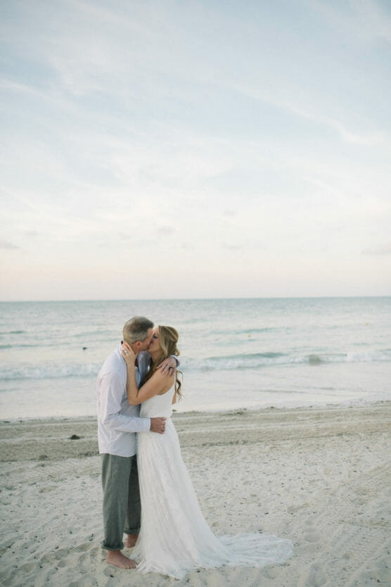 A couple dressed in wedding attire kisses on a sandy beach at sunset, with gentle waves and a soft sky in the background. by Justin Salem Meyer