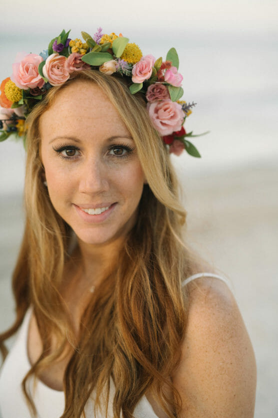A woman with long, wavy auburn hair wears a colorful flower crown and a white dress, smiling softly while standing on a sandy beach with blurred water in the background. by Justin Salem Meyer