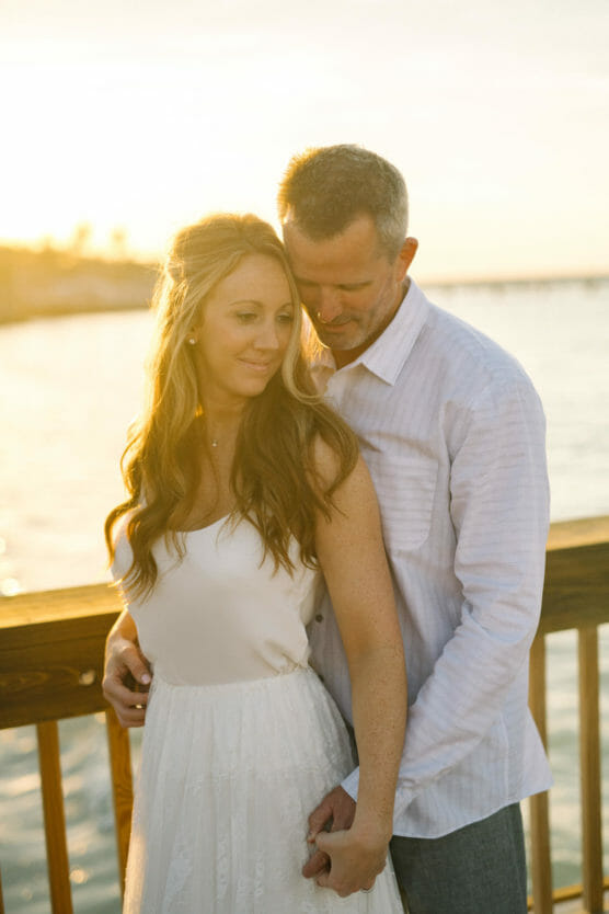 A couple stands on a wooden pier at sunset. The woman in a white dress smiles slightly while the man in a white shirt embraces her from behind, both looking content with the sunlit water in the background. by Justin Salem Meyer