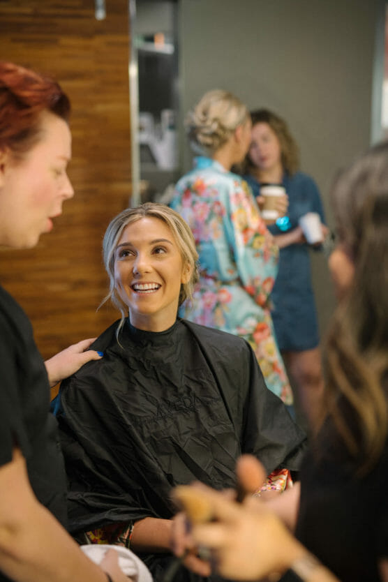 A smiling woman wearing a black cape sits in a salon chair while talking to two people. In the background, two women stand and chat, one in a floral robe holding a cup. The setting appears to be a lively salon or getting-ready area. by Justin Salem Meyer