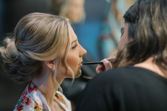 A woman with blonde hair styled in an updo is having her makeup done by another person, who is applying lipstick with a brush. The woman wears a floral robe and looks ahead calmly. by Justin Salem Meyer