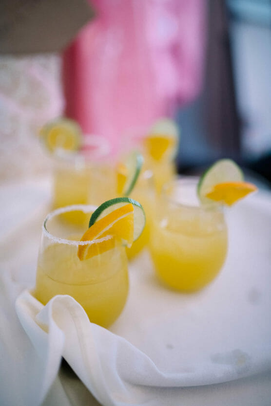 Five glasses of yellow cocktail garnished with lime and orange slices are arranged on a white cloth, with a blurred pink background. by Justin Salem Meyer