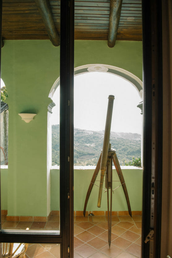 A large brass telescope on a tripod stands on a tiled balcony, framed by an arched opening and green walls, overlooking a hilly, sunlit landscape in the distance. by Justin Salem Meyer