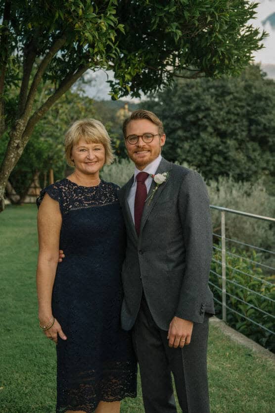 A woman in a navy lace dress and a man in a grey suit with a red tie and boutonniere stand smiling together outdoors near a railing, with trees and greenery in the background. by Justin Salem Meyer