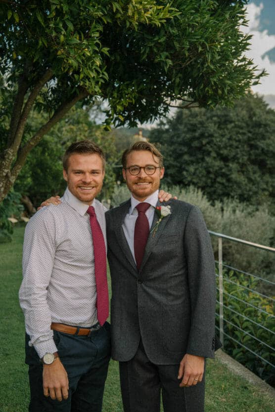 Two men stand close together outside, smiling at the camera. One wears a grey suit with a red tie and boutonniere, while the other wears a white shirt and red tie. They are under a tree, with greenery in the background. by Justin Salem Meyer