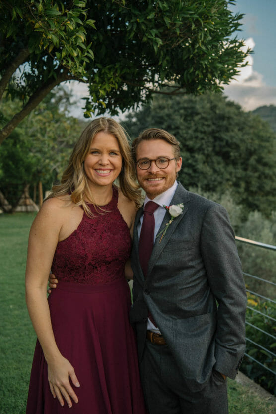 A smiling woman in a sleeveless burgundy dress stands beside a man in a gray suit and burgundy tie with a boutonnière, posing outdoors near greenery and a metal railing. by Justin Salem Meyer