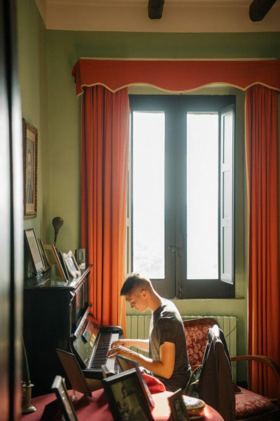 A young man sits at an upright piano, playing music in a sunlit room with green walls and orange curtains. Framed photos and decorations are visible on the piano and table nearby. by Justin Salem Meyer